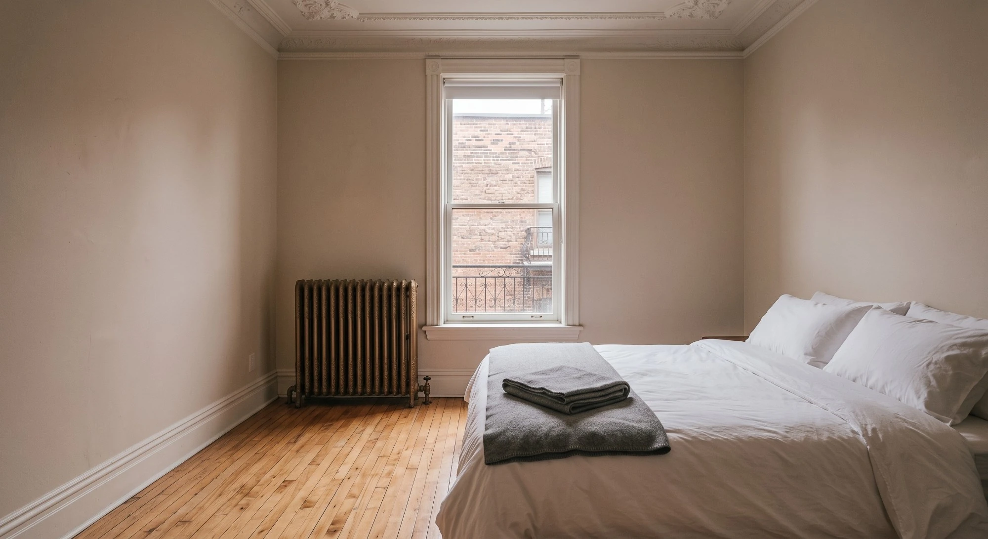 Illustration: a bedroom with hardwood floors, a cast-iron radiator and a made bed with a folded blanket