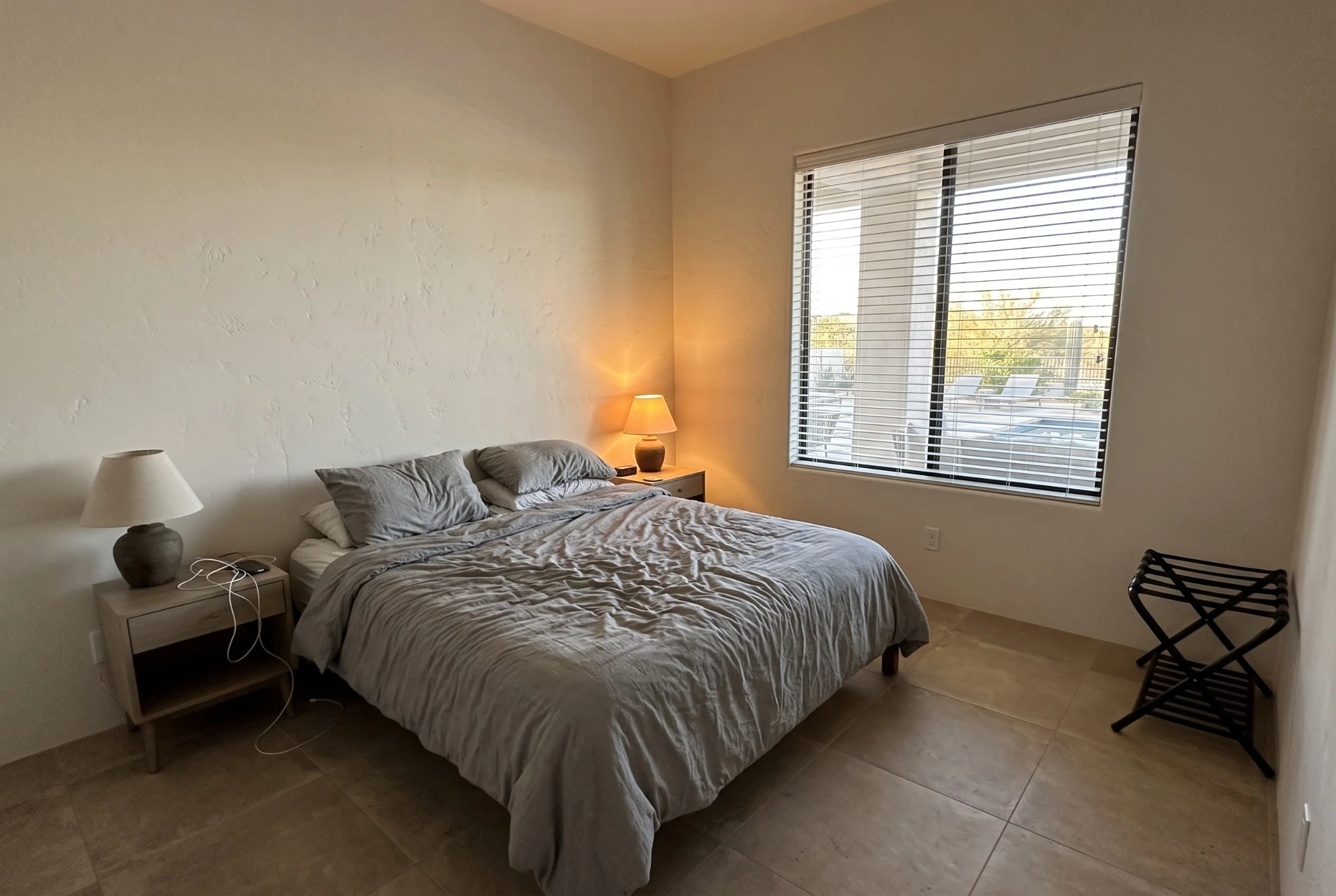 Illustration, before: a guest bedroom with an unmade grey duvet, flat pillows, cables on the nightstand and a luggage rack